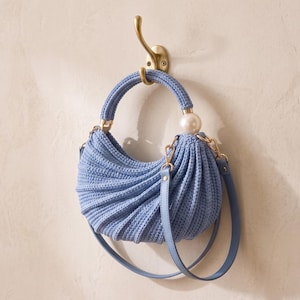 May include: A crescent-shaped light blue crochet handbag featuring a textured ribbed pattern, a rounded handle, and a large white spherical bead accent. The bag includes a matching light blue shoulder strap attached with gold-toned metal hardware.