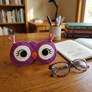 May include: A purple crocheted owl mask featuring large white circular eyes with black pupils and long eyelashes, an orange beak, and small tufts of multicolored yarn on each side. The mask is positioned on a wooden surface next to eyeglasses.