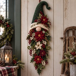 May include: A vertical wall hanging featuring a crocheted gnome with a tall dark green pointed hat, a cream-colored brim, and a round nose. The gnome is adorned with crocheted red and white poinsettia flowers, green leaves, pinecones, and red berries.
