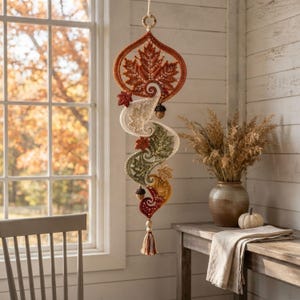 May include: A hanging crochet wall decoration featuring a large orange maple leaf at the top, followed by a cascading spiral of cream, sage green, and mustard yellow leaves. The piece is accented with small brown acorns and a bottom tassel.