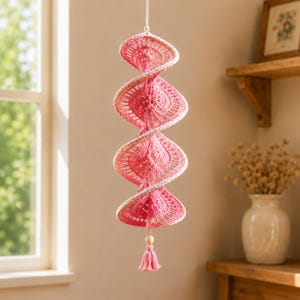 May include: A hanging spiral ornament crafted from pink and white crochet thread. The structure features a repeating openwork pattern that twists vertically, ending in a small wooden bead and a matching pink crochet tassel at the bottom.