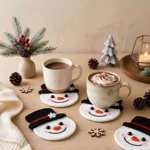 May include: Four round crochet coasters shaped like snowman faces, each featuring a black top hat with a red band, a small orange carrot nose, black eyes, and pink cheeks. Two ceramic mugs filled with hot beverages sit on top of two coasters.