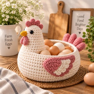May include: A cream-coloured crochet chicken basket featuring a pink comb, wattle, and wing accents. The chicken-shaped container holds several brown and white eggs. The body is crafted from textured yarn stitches, with black bead eyes.