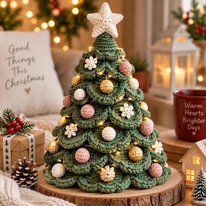 Puede incluir: Un árbol de Navidad de ganchillo verde con una estrella blanca en la punta, decorado con pequeñas esferas de ganchillo blancas, rosas y doradas y adornos de copos de nieve blancos. El árbol está iluminado con luces y sobre una base de madera.