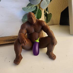 May include: A brown 3D printed figurine of a muscular person in a seated position with legs spread apart. A purple eggplant emoji is positioned over the pelvic area of the figure. The figurine has a smooth, solid brown finish and defined muscles.