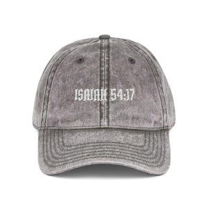 May include: A charcoal grey acid-wash baseball cap featuring the embroidered white text Isaiah 54:17 centred on the front panel. The cap has a curved brim, a button on the crown, and stitched eyelets for ventilation, providing a structured look.