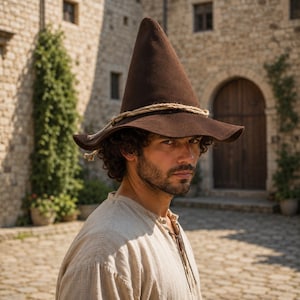 May include: A dark brown pointed hat with a wide, slightly curved brim and a thick, light-coloured rope tied around the base of the crown. The hat features a smooth, felt-like texture and is worn by a person with dark, curly hair and a beard.