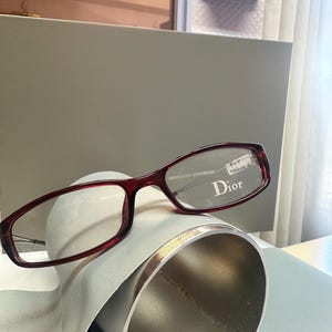 May include: Dark red rectangular eyeglasses with a thin frame. The right lens features the white text Dior and a small white sticker with the text PVP 199.00 €. The glasses have a sleek design with thin temples and a classic bridge structure.