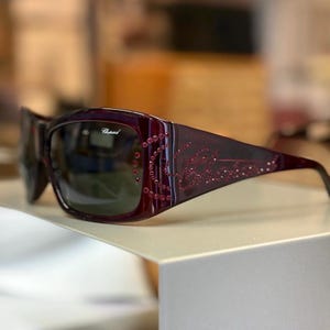 May include: Dark burgundy sunglasses featuring a rectangular frame with dark tinted lenses. The temples are embellished with small red crystals arranged in a swirling pattern and the Chopard brand name is visible on the lens and temple.