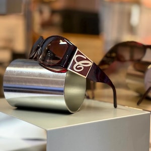 May include: Dark burgundy sunglasses featuring a rectangular frame and gradient lenses. The temples are adorned with a gold-toned metal plaque displaying a cursive letter C logo embellished with small, clear, sparkling crystals.