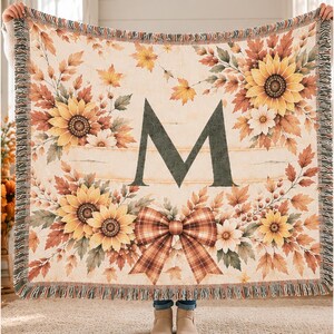 May include: A woven throw blanket featuring a large dark green letter M in the center, surrounded by yellow sunflowers, autumn leaves, and a plaid bow. The blanket has a cream-colored background and a fringed edge on all four sides.