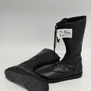 May include: A pair of black mid-calf boots with a lace-up front closure and a flat, textured rubber sole. A white tag attached to the laces features the handwritten text "The Deal DIGGER" in black ink. The boots have a smooth, matte finish.