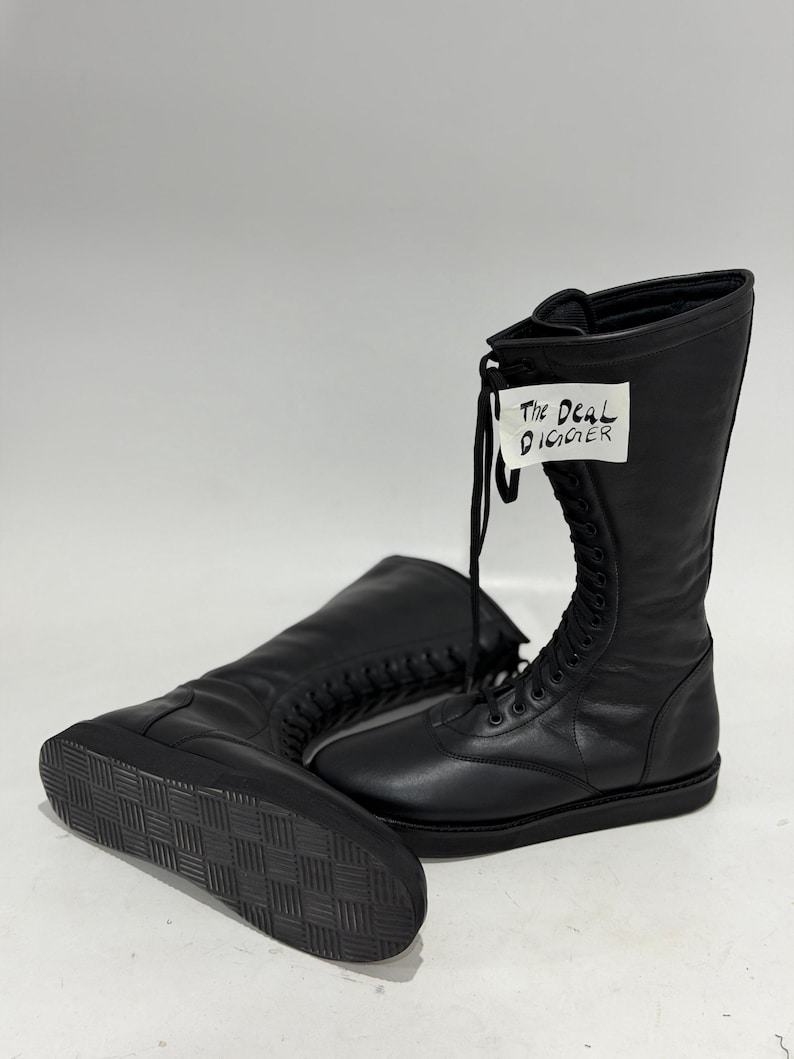 May include: A pair of black mid-calf boots with a lace-up front closure and a flat, textured rubber sole. A white tag attached to the laces features the handwritten text "The Deal DIGGER" in black ink. The boots have a smooth, matte finish.