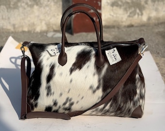 May include: A cowhide duffel bag featuring a black and white spotted pattern with dark brown leather handles, trim, and a detachable shoulder strap. Two white tags are attached to the top, each printed with the text The Deal Digger.