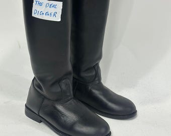 May include: A pair of tall black leather boots with a smooth finish and rounded toes. A white rectangular tag is attached to the side of the left boot with the text THE DEAL DIGGER written in blue ink. The boots feature a flat sole and a structured shaft.