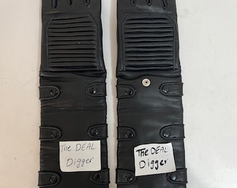 May include: A pair of black leather gloves featuring ribbed detailing on the back of the hand, multiple snap button closures along the forearm, and a white paper label on each glove with the handwritten text The DEAL Digger in black ink.