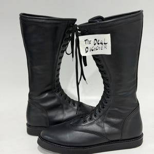 May include: A pair of black knee-high lace-up boots with a smooth finish, rounded toes, and flat black soles. The boots feature long black laces and a white tag attached to the front with the handwritten text The Deal Digger in black ink.