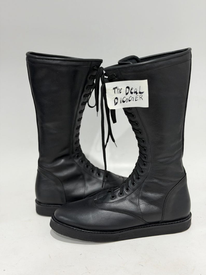 May include: A pair of black knee-high lace-up boots with a smooth finish, rounded toes, and flat black soles. The boots feature long black laces and a white tag attached to the front with the handwritten text The Deal Digger in black ink.