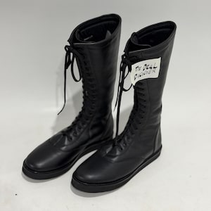 May include: A pair of black knee-high lace-up boots with a smooth finish, rounded toes, and flat black soles. The boots feature long black laces and a white tag attached to the right boot with the handwritten text The Deal Digger in black ink.