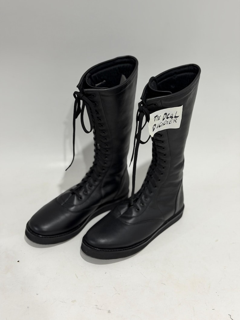 May include: A pair of black knee-high lace-up boots with a smooth finish, rounded toes, and flat black soles. The boots feature long black laces and a white tag attached to the right boot with the handwritten text The Deal Digger in black ink.