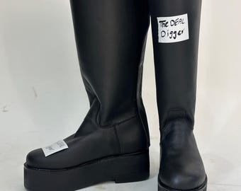 May include: A pair of black knee-high boots with thick platform soles. The boots feature a smooth matte finish and a rounded toe. A white rectangular label with the black text The DEAL Digger is attached to the side of each boot shaft.