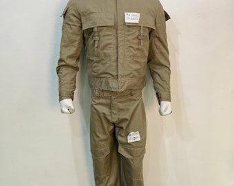 May include: A tan utility jacket and matching trousers set worn by a mannequin. The jacket features a high collar, two chest pockets with flaps, and vertical straps. The trousers include large cargo-style pockets. A white tag reads The Deal Digger.