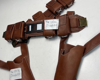 May include: A brown leather utility belt featuring multiple attached pouches, a black central buckle component, and two integrated holsters. The belt includes white stitched accents and two white paper labels with the text The DEAL DIGGER.