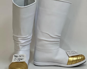 May include: A pair of tall white boots featuring a metallic gold rounded toe cap with horizontal stitching. Each boot has a white tag attached to the front displaying the text The DEAL DIGGER in black ink. The boots have a slouchy shaft design.