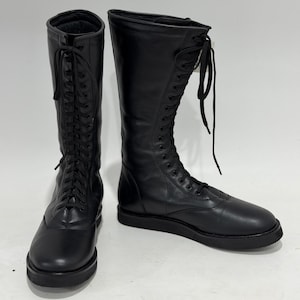 May include: A pair of black mid-calf boots featuring a smooth leather-like finish, a rounded toe, and a full-length front lace-up closure with matching black laces. The boots have a flat, thick black sole and a structured shaft that rises to the mid-calf.