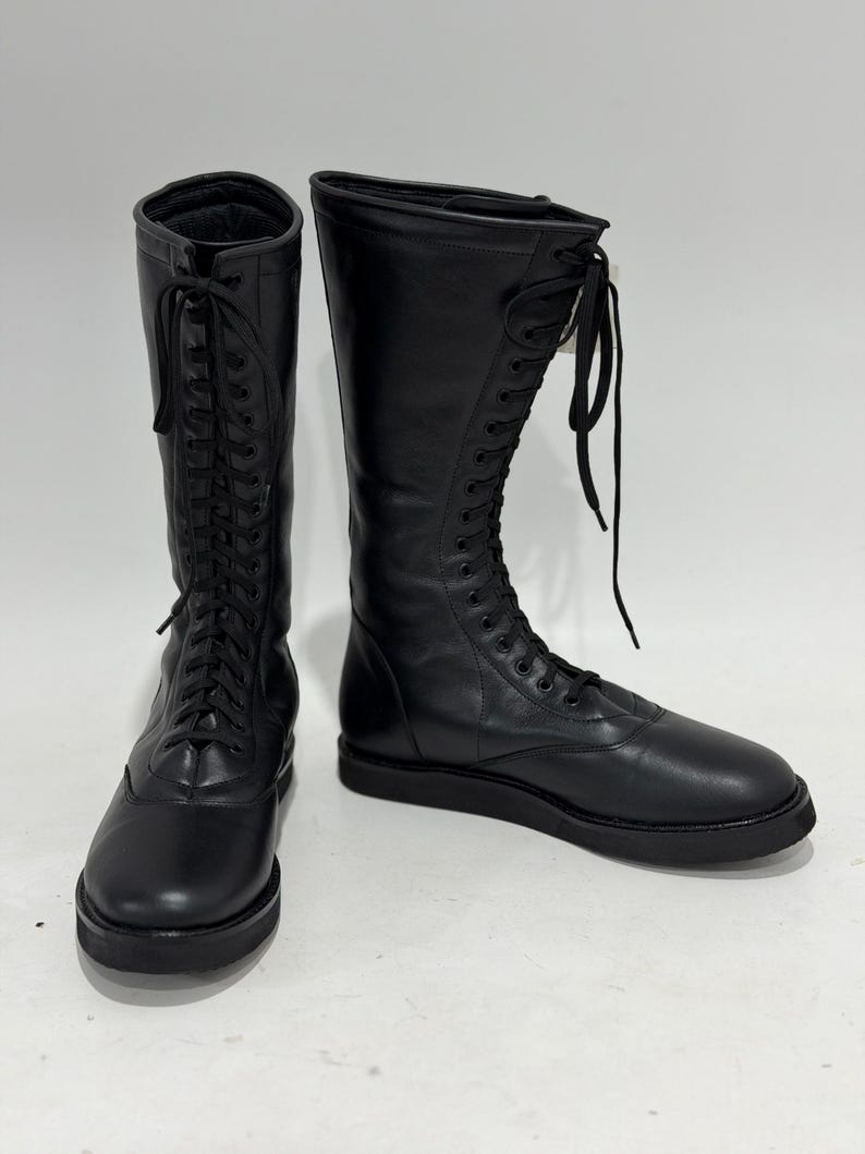 May include: A pair of black mid-calf boots featuring a smooth leather-like finish, a rounded toe, and a full-length front lace-up closure with matching black laces. The boots have a flat, thick black sole and a structured shaft that rises to the mid-calf.