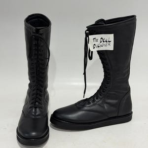 May include: A pair of black leather mid-calf boots featuring a rounded toe, a flat sole, and a full-length lace-up front with black laces. One boot displays a white tag with the handwritten text The Deal Digger attached to the upper shaft.