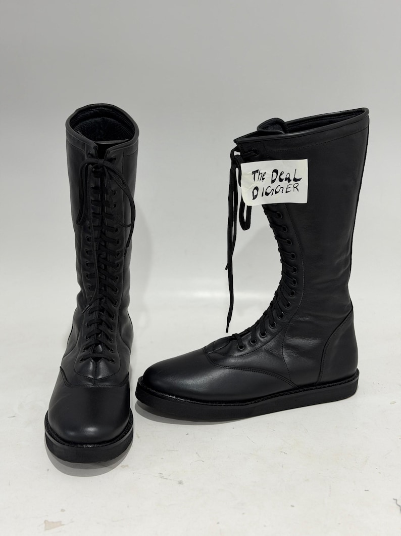 May include: A pair of black leather mid-calf boots featuring a rounded toe, a flat sole, and a full-length lace-up front with black laces. One boot displays a white tag with the handwritten text The Deal Digger attached to the upper shaft.