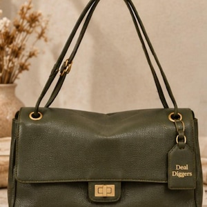 May include: An olive green shoulder bag with a textured finish, featuring a gold-toned rectangular turn-lock closure, matching gold hardware, and a hanging luggage tag embossed with the text Deal Diggers. The bag has two adjustable shoulder straps.