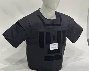 May include: A black short-sleeved tactical vest featuring multiple rectangular hook-and-loop fastener strips on the chest and shoulders. A small white rectangular label with the blue text THE DEAL DIGGER is attached to the center of the chest.