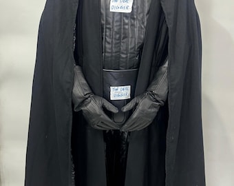 May include: A black costume featuring a full-head mask, a long cape, and gloves. The chest and belt area each display a white rectangular label with the handwritten text THE DEAL DIGGER. The outfit includes a quilted black vest and dark attire.