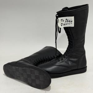 May include: A pair of black mid-calf boots with a lace-up front closure and a flat, textured rubber sole. A white tag attached to the laces features the handwritten text "The Deal Digger" in black ink. The boots have a smooth, matte finish.