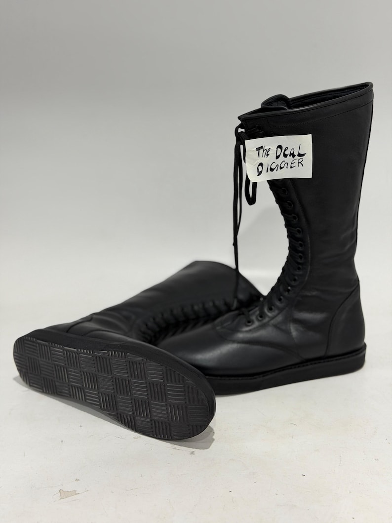 May include: A pair of black mid-calf boots with a lace-up front closure and a flat, textured rubber sole. A white tag attached to the laces features the handwritten text "The Deal Digger" in black ink. The boots have a smooth, matte finish.