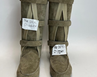 May include: A pair of olive green suede knee-high boots featuring a moccasin-style toe, three horizontal straps across the shaft, and vertical accent strips. Each boot displays a white paper tag with the handwritten text The DEAL DIGGER.