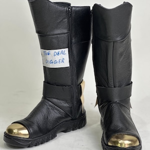 May include: A pair of black knee-high boots with a textured leather finish, featuring a metallic gold-toned toe cap, a side zip closure, and a horizontal strap across the mid-foot. A white label with the text THE DEAL DIGGER is attached.