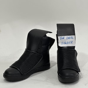 May include: A pair of black high-top trainers featuring a smooth leather toe and a ribbed, padded mid-foot strap. The shoes have a tall, structured collar and a flat rubber sole. A white paper tag with the text THE DEAL DIGGER is attached.