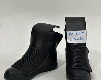 May include: A pair of black high-top sneakers featuring a smooth leather toe and a ribbed, padded mid-foot strap. The shoes have a tall, structured collar and a flat rubber sole. A white paper tag with the text THE DEAL DIGGER is attached.