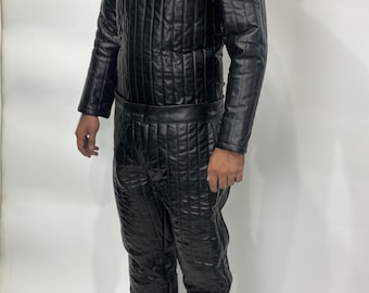 May include: A black quilted leather-look jumpsuit featuring a high neck, long sleeves, and vertical stitching throughout. A small white rectangular tag with the text "THE DEAL DIGGER" is attached to the chest area of the garment.