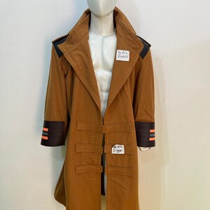 Può includere: Un cappotto lungo color marrone chiaro con colletto ampio, dettagli neri sulle spalle e polsini marrone scuro con due strisce arancioni orizzontali. Il cappotto presenta tre chiusure a linguetta orizzontali e due etichette The DEAL DIGGER.