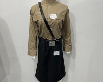 May include: A tan long-sleeved jacket with a high collar and two chest pockets, paired with a black skirt and a black belt featuring a silver buckle and a diagonal strap. Two white labels with the text The DEAL Digger are attached to the outfit.