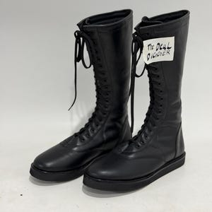 May include: A pair of black knee-high lace-up boots with a flat sole and rounded toe. The boots feature long black laces and a white tag attached to the upper shaft with the handwritten text The Deal Digger in black ink.