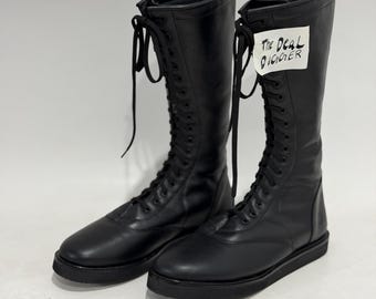 May include: A pair of black knee-high lace-up boots with a flat sole and rounded toe. The boots feature long black laces and a white tag attached to the upper shaft with the handwritten text The Deal Digger in black ink.
