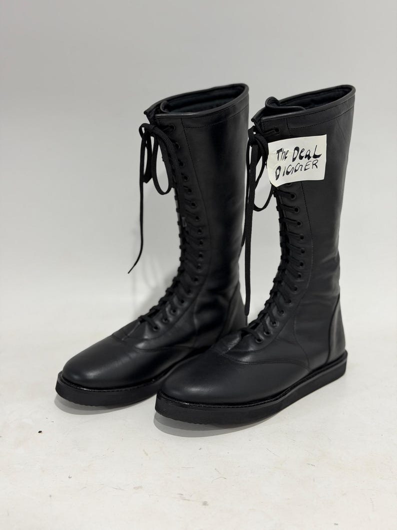 May include: A pair of black knee-high lace-up boots with a flat sole and rounded toe. The boots feature long black laces and a white tag attached to the upper shaft with the handwritten text The Deal Digger in black ink.