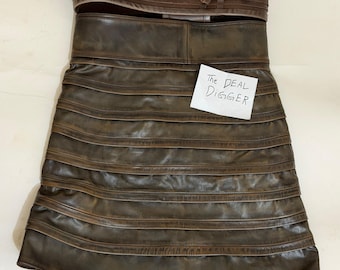 May include: A dark brown leather skirt featuring horizontal layered panels and a wide waistband with integrated loops. A white rectangular tag with the handwritten text The DEAL DIGGER is attached to the front of the skirt, which has a smooth finish.