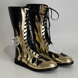 May include: A pair of tall, black boots featuring metallic gold flame-shaped overlays, black laces, and a black sole. Each boot has a white tag attached with the text "The DEAL Digger" printed in black ink, visible on the side and front.