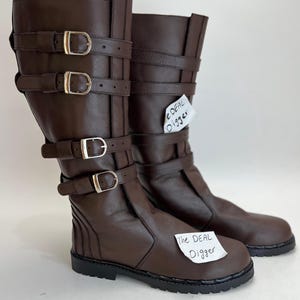 May include: A pair of dark brown mid-calf boots featuring four horizontal straps with silver-toned metal buckles on the outer side. The boots have a rounded toe, a thick black lug sole, and a paper tag attached with the text The DEAL Digger.